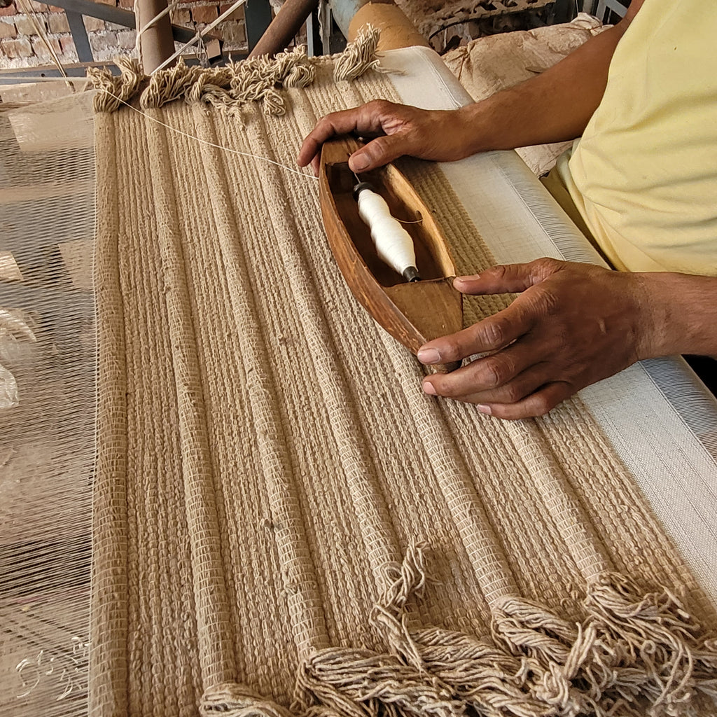 Weaver's hands passing a wooden shuttle across a clay-brown Myrobalan yoga mat on the handloom