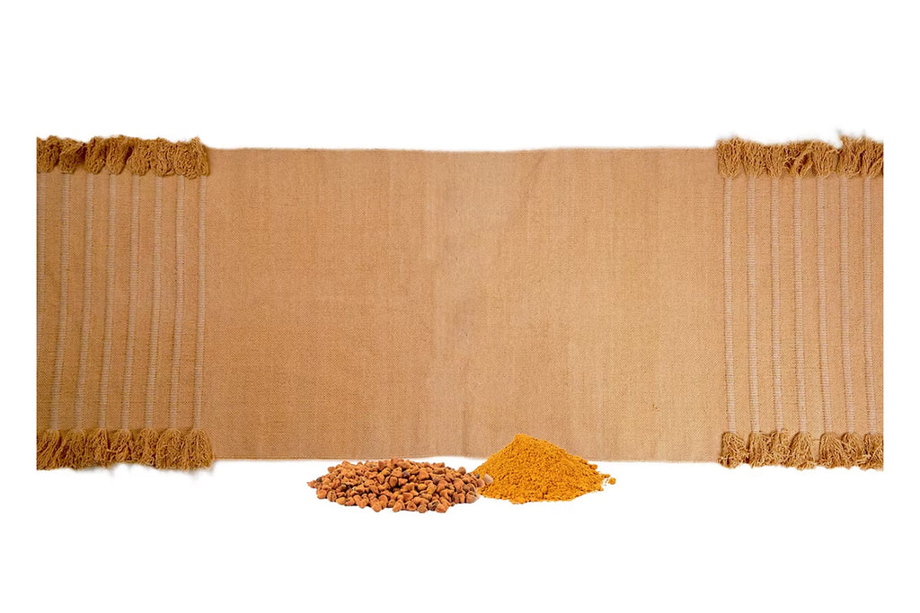 Ayurvastra yoga rug in warm annatto red-orange displayed with annatto root and powdered annatto herb, showcasing the natural Ayurveda plant dyes used in the herbal textile process